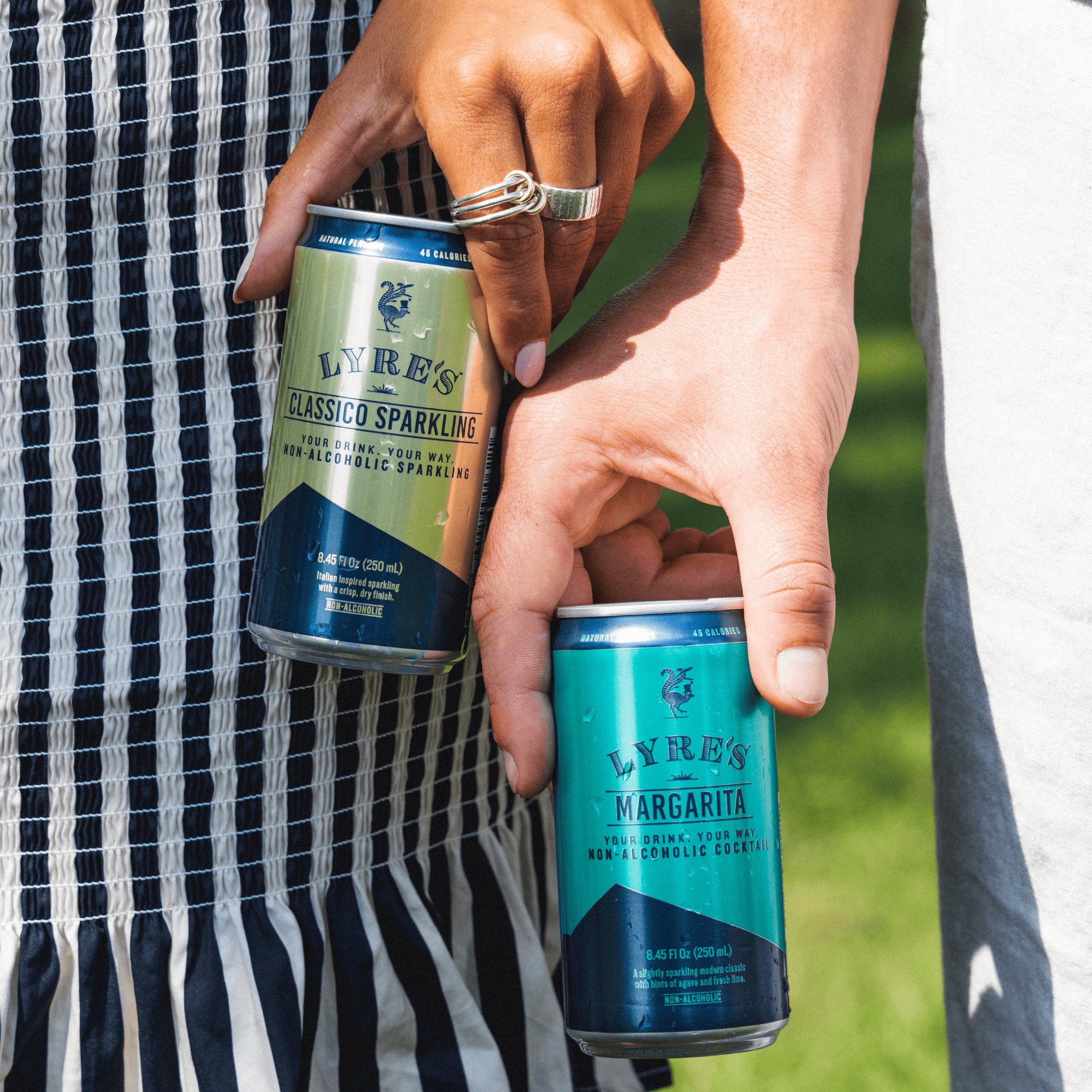 Two hands holding Lyres non-alcoholic sparkling and margarita cocktails in a can against a grassy background.