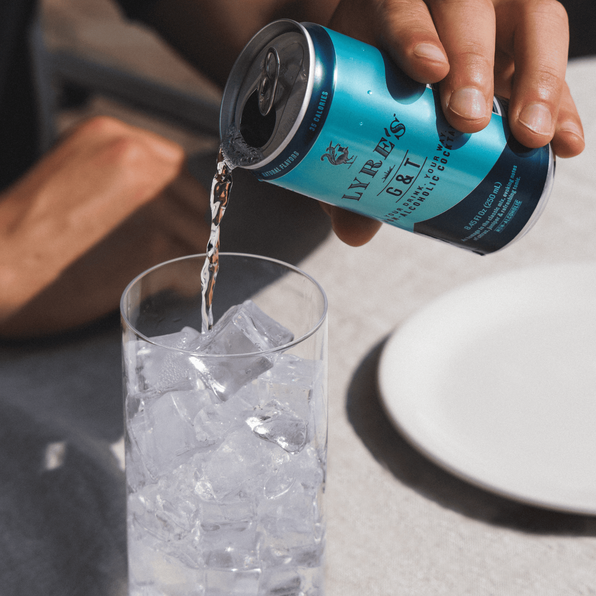 Person pouring a can of Lyre's G&T non alcoholic cocktail into a glass with ice on a light surface.