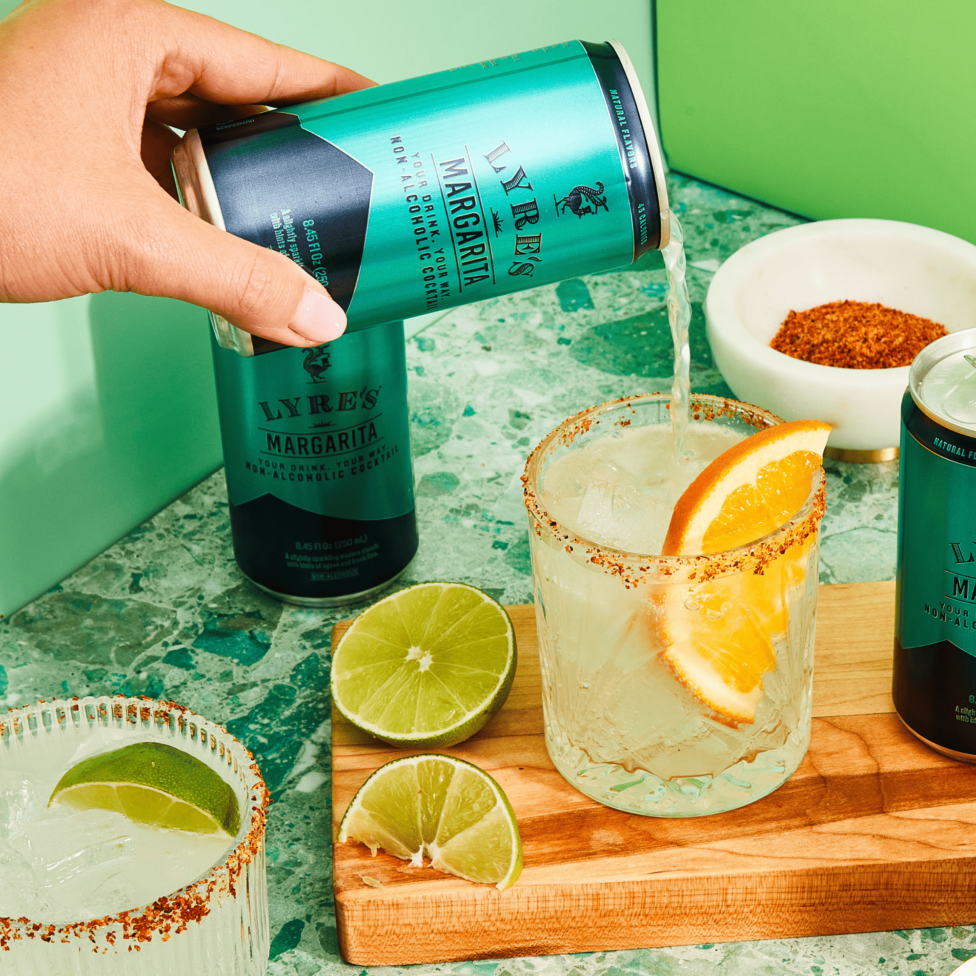 Hand pouring Lyne's non alcoholic Margarita into a glass with lime and orange slices on a green countertop.