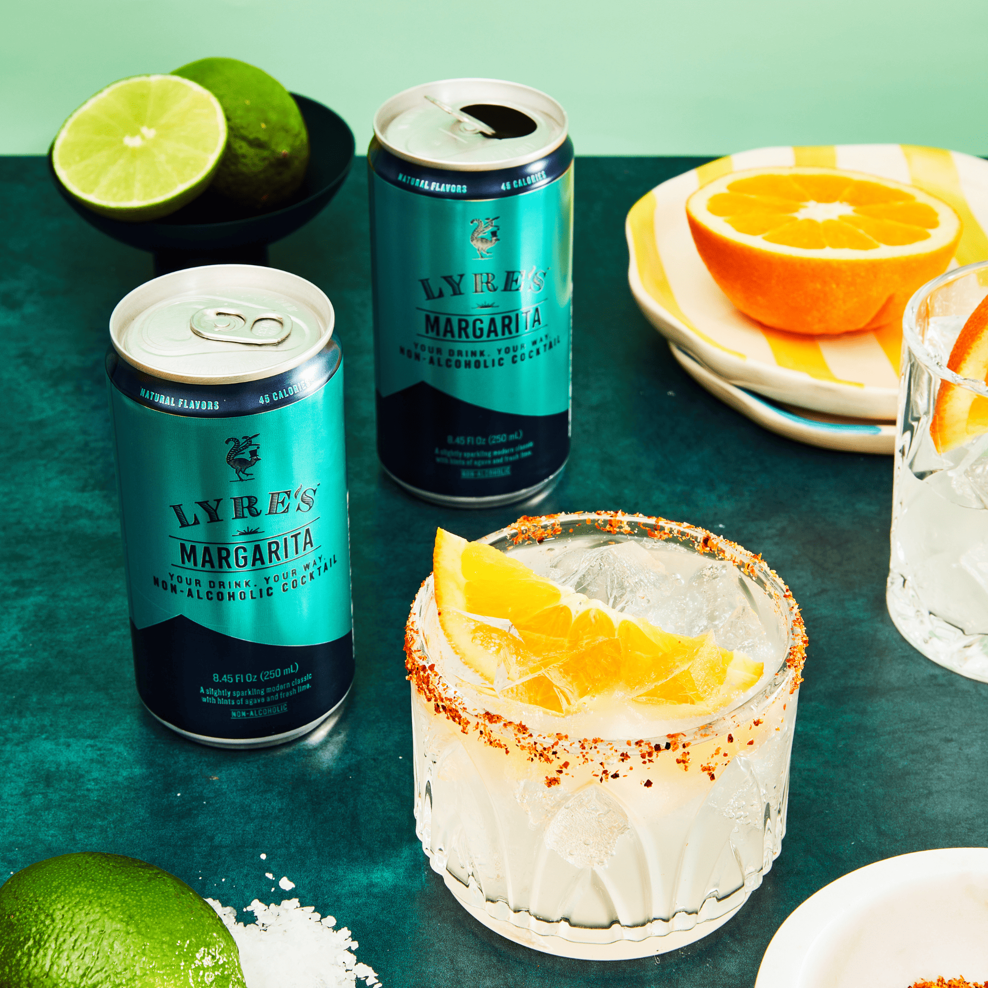 Two cans of Lyre's Margarita non alcoholic cocktail with a cocktail garnished with orange on a green table.