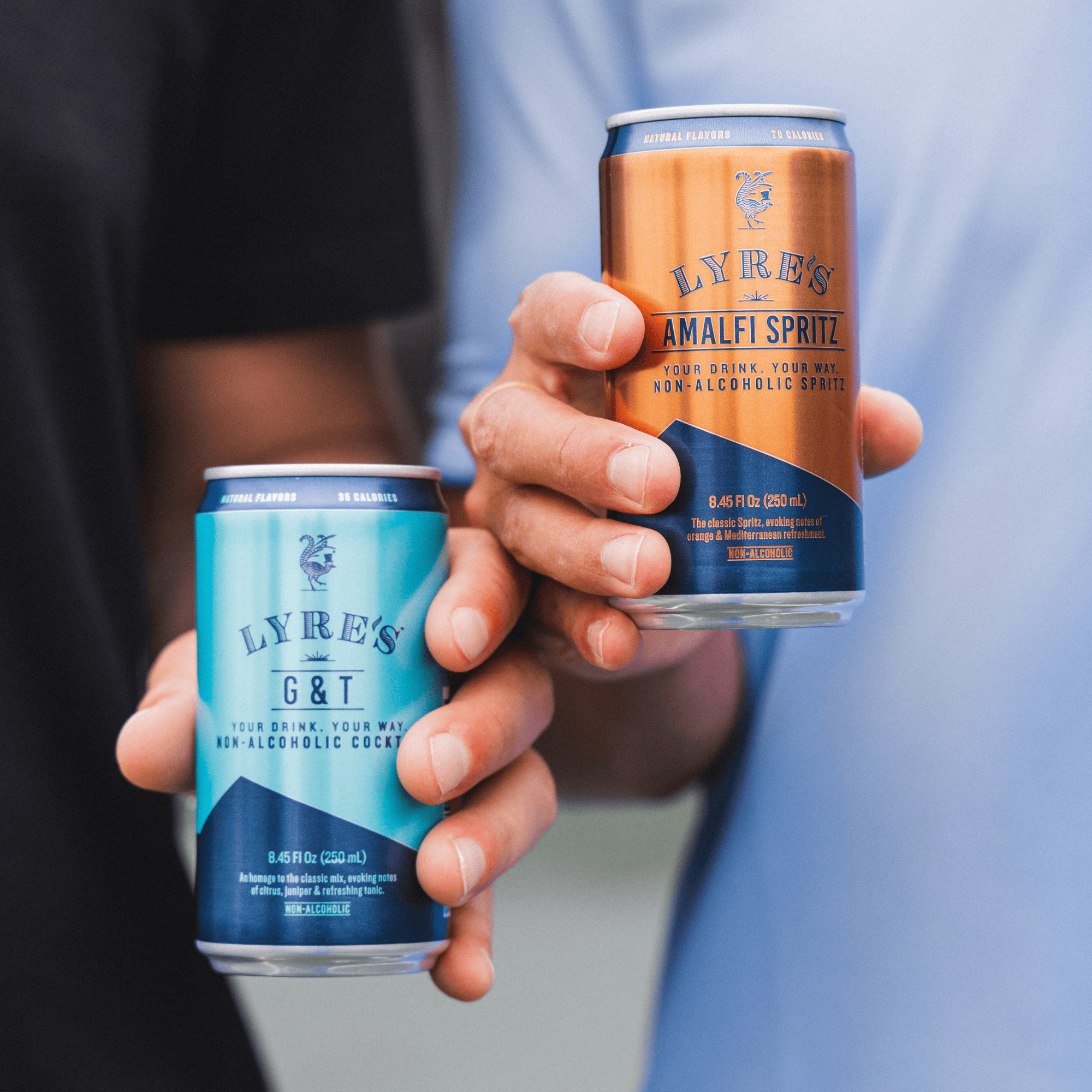 Two cans of Lyre's non-alcoholic cocktails held by a person against a blurred background