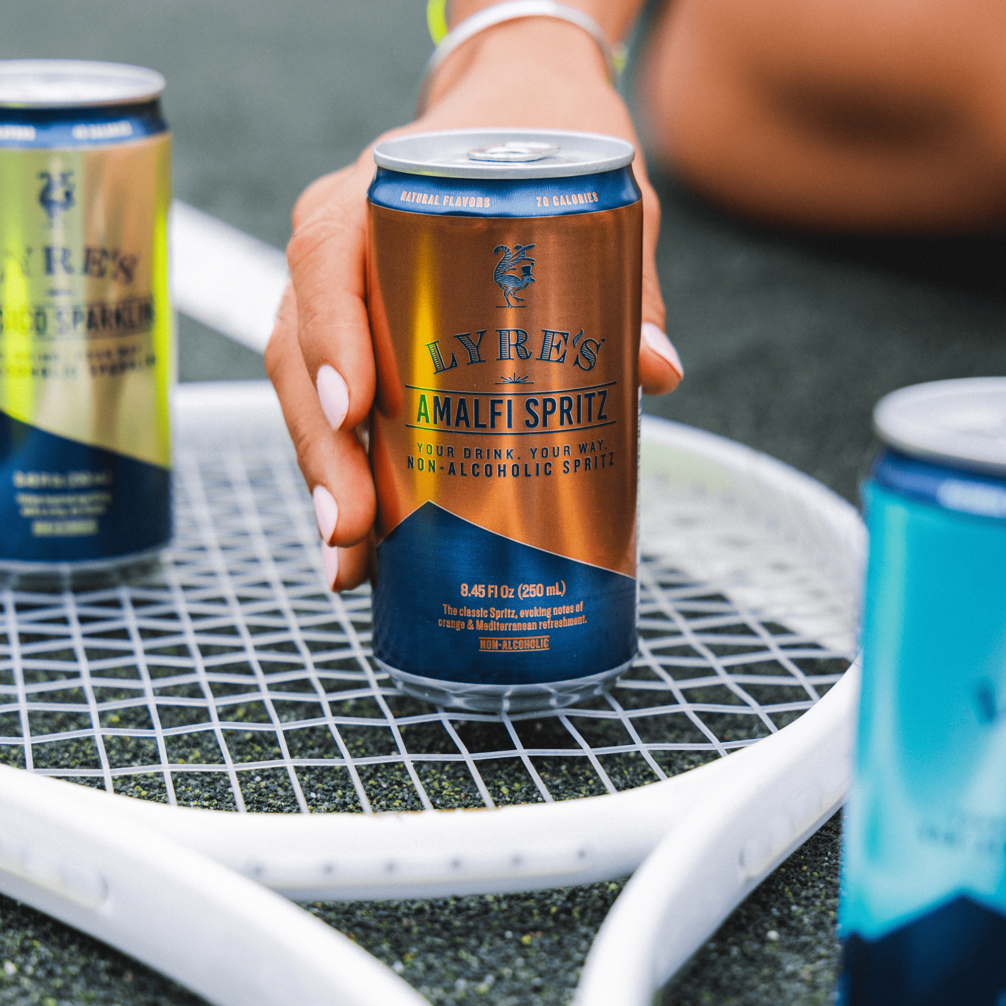 Hand holding a can of Lyre's Amalfi Spritz non alcoholic cocktail on a tennis racket with other cans in the background.