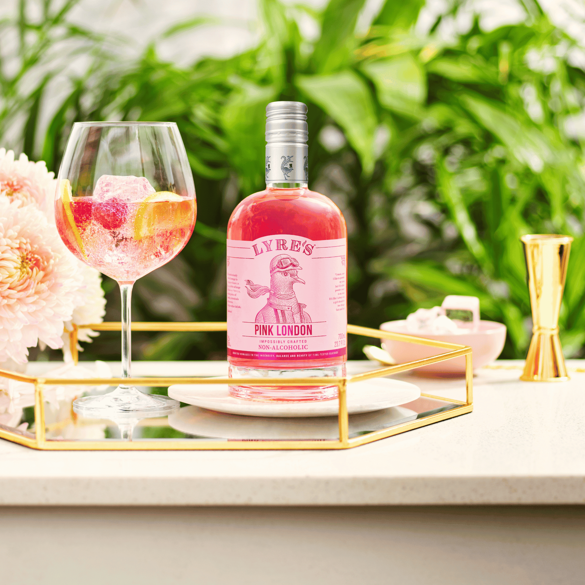 Bottle of Lyre's Pink London with a glass of pink spritz cocktail on a decorative stand with flowers and greenery in the background.