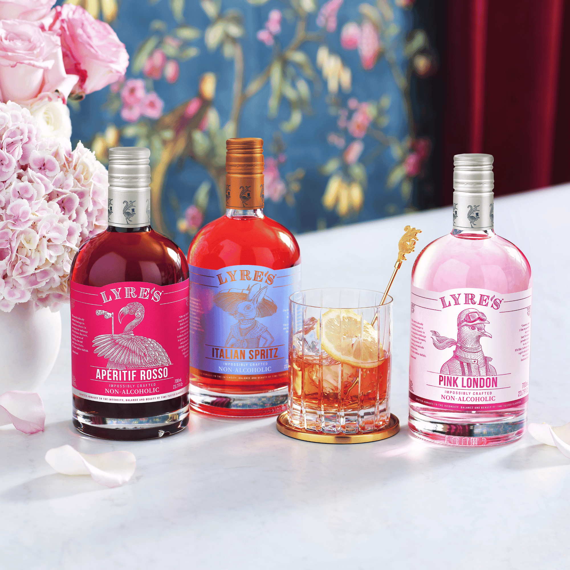 Three bottles of Lyre's Aperitif Rosso, Italian Spritz and Pink London on a table with a floral arrangement in the background.