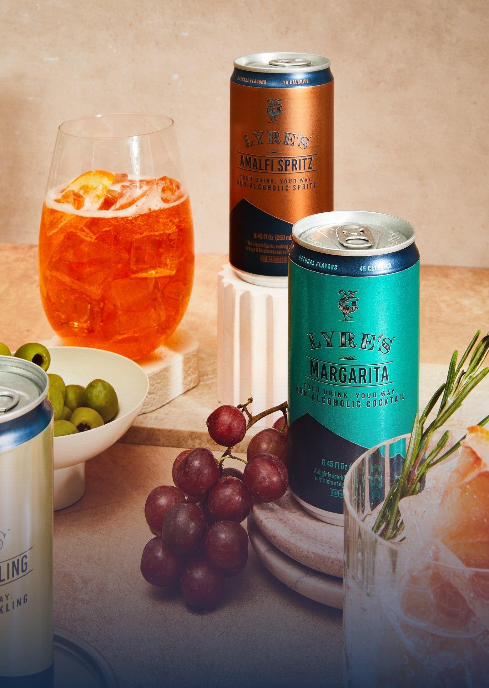 Two cans of Lyre's non alcoholic cocktail Mand Amalfi Spritz on a table with a glass of orange cocktail and bowl of olives.